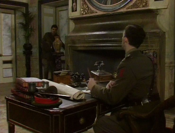 Blackadder walking into Melchett's office, with the fireplace and circular portrait visible