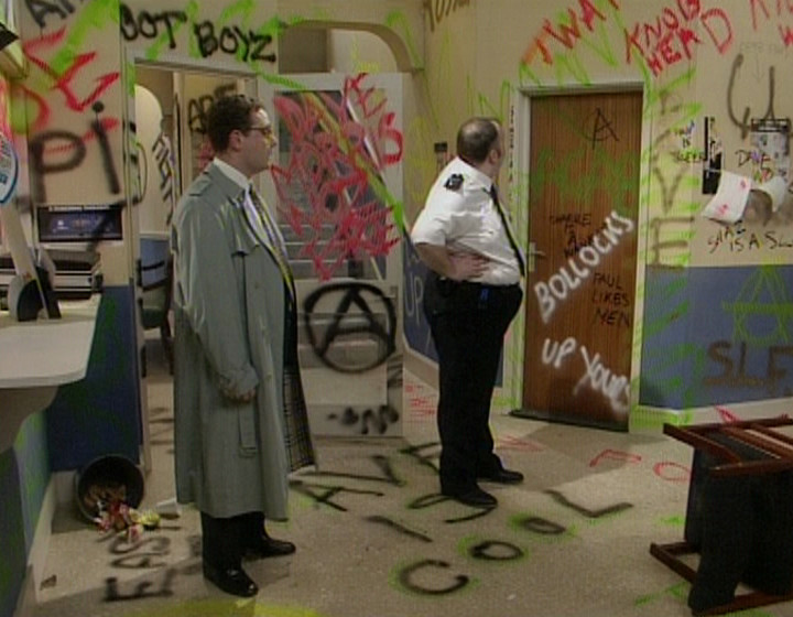 John Thomson as a respectable man, standing in a police station covered in graffiti