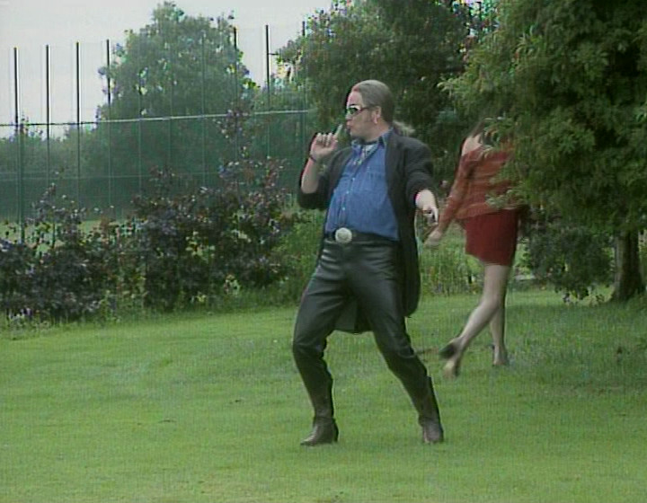 Charlie Higson dressed in tight leather trousers, blowing the smoke from a fake gun made from his fingers