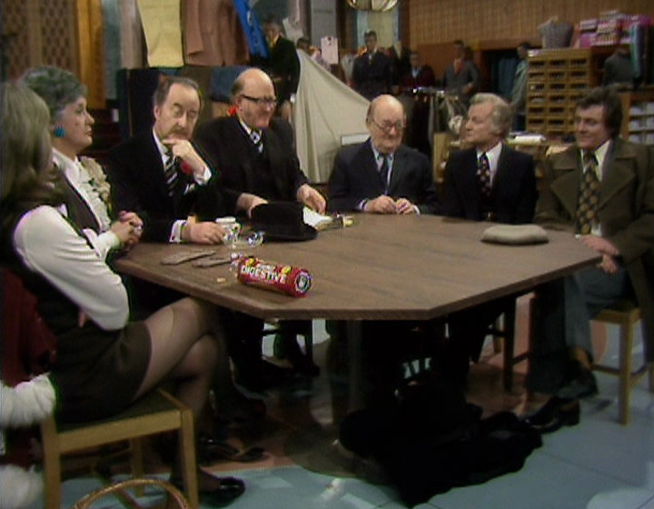 The Are You Being Served? gang around a table on the shop floor, with a gap around one side, from a slightly different angle